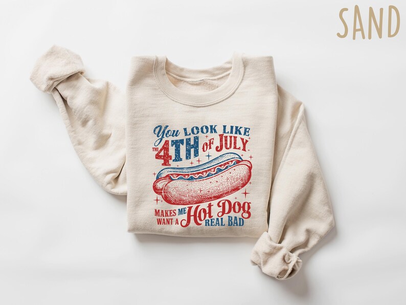 Op de afbeelding: Zandkleurige sweatshirt met een afbeelding van een hotdog en de tekst "You look like the 4th of July makes me want a hot dog real bad" in rood en blauw. De sweatshirt is opgevouwen.