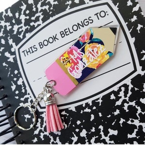 May include: A pencil-shaped keychain with a floral design and the name "Ms. Gonzalez" in white lettering. The keychain has a pink eraser, a silver chain, and a pink tassel. The background is a notebook with the text "THIS BOOK BELONGS TO:"