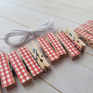 May include: A set of wooden clothespins with red and white gingham patterned tops, arranged with a white rope on a light wood surface. The clothespins are made of natural wood and have metal springs. The gingham pattern adds a decorative touch.