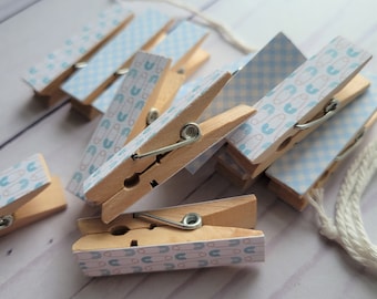 May include: A set of wooden clothespins with a blue and white gingham pattern and a blue and white pattern of safety pins. The clothespins are tied together with a white string.