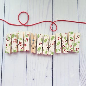 May include: A collection of wooden clothespins with a festive holly and berry pattern. The clothespins are arranged in a row, connected by a red string, creating a decorative element. The background is a light wood surface.