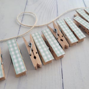 May include: Wooden clothespins with a light green and white gingham pattern are attached to a natural twine. The clothespins are arranged in a row, and the background is a light-colored wooden surface.