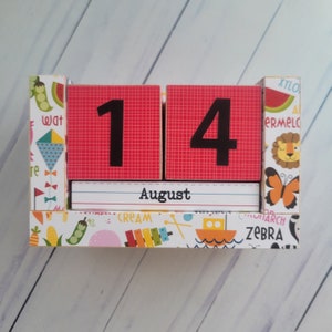 May include: A red and white wooden calendar block set with the date August 14th. The calendar blocks have a colorful patterned background with images of animals, food, and other objects.