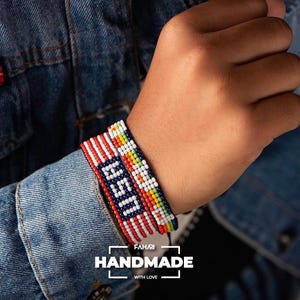 May include: Two beaded bracelets on a wrist. One bracelet is red, white, and blue with a white "USA" design. The other bracelet is multi-colored with red, orange, yellow, green, and white beads. The bracelets are handmade.