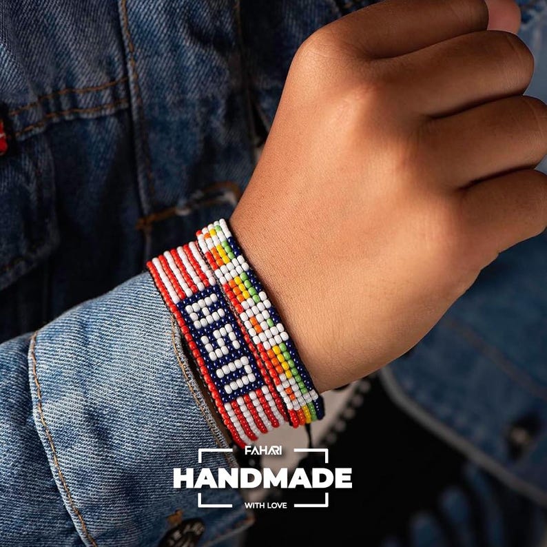 May include: Two beaded bracelets on a wrist. One bracelet is red, white, and blue with a white "USA" design. The other bracelet is multi-colored with red, orange, yellow, green, and white beads. The bracelets are handmade.