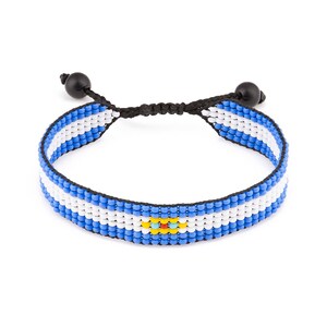 May include: A woven bracelet featuring the colors of the Argentinian flag: light blue, white, and blue. The bracelet has a black adjustable cord with black beads at each end. The center of the bracelet features a small emblem.