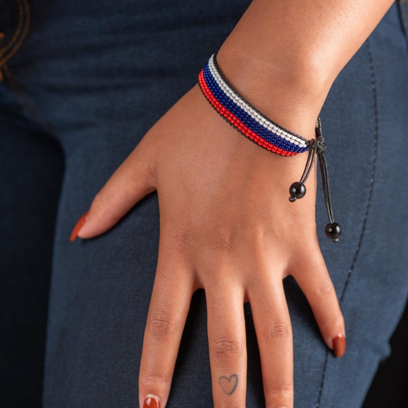 Op de afbeelding: Een close-up van iemands pols met een kralenarmband met rode, witte en blauwe kralen. De armband is vastgebonden met een zwart koord en heeft zwarte kralen aan de uiteinden.