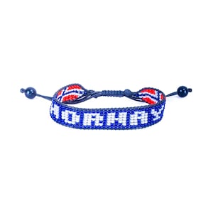 May include: A woven beaded bracelet featuring the word "NORWAY" in white beads against a blue background. The design includes red and white accents along the edges, with dark blue cord ties ending in single dark blue beads on each side.
