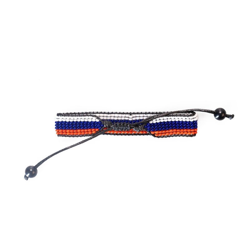 Op de afbeelding: Een kralen armband met oranje, witte en blauwe strepen. De armband is vastgebonden met een zwart koord en heeft zwarte kralen aan de uiteinden.