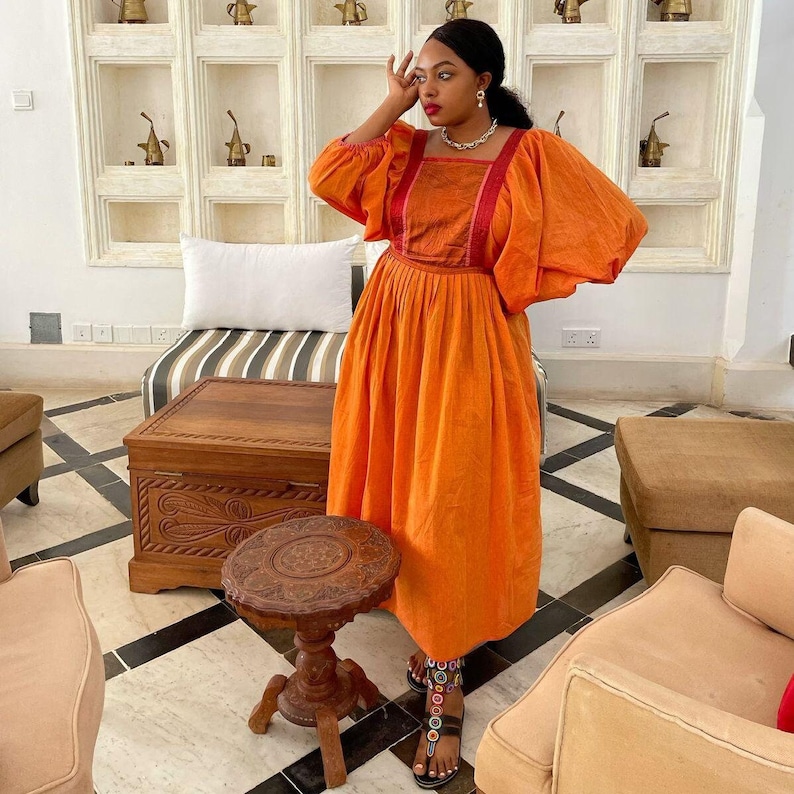 Puede incluir: Una mujer con un vestido largo de color naranja con ribetes rojos y blancos. Est&aacute; de pie en una habitaci&oacute;n con un cofre de madera y una mesa peque&ntilde;a. La mujer lleva sandalias con cuentas de colores.