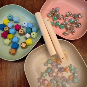 以下が含まれることがあります： 3つの別々のボウルに、カラフルな木製ビーズ、キラキラの透明ガラスビーズ、パステルカラーのプラスチックビーズが詰められています。木製ダボ2本も含まれています。