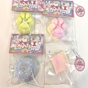 Könnte beinhalten: Vier bunte, weiche Spielzeuge in einzelnen durchsichtigen Plastiktüten mit einem kleinen Etikett, auf dem in chinesischen Schriftzeichen "Xiao Xiao" steht. Die Spielzeuge sind in Form einer Pfote, eines Donuts und zweier Rechtecke. Die Pfote ist grün, der Donut ist lila mit weißen Streuseln und die Rechtecke sind rosa und gelb. Die Beutel sind mit Maßen in Zentimetern beschriftet.