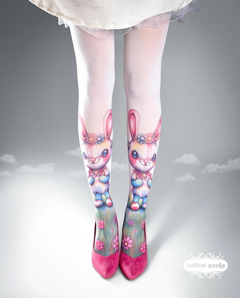 May include: White tights with a colorful bunny design. The tights feature a cartoon bunny with a floral crown and a blue and pink outfit. The bunny is standing on a grassy hill with pink flowers. The model is wearing pink heels.