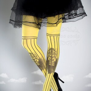 Puede incluir: Mallas amarillas con rayas negras y un estampado gr&aacute;fico de un gato con un gorro de marinero y una camisa a rayas. El gato est&aacute; detr&aacute;s de las rejas.