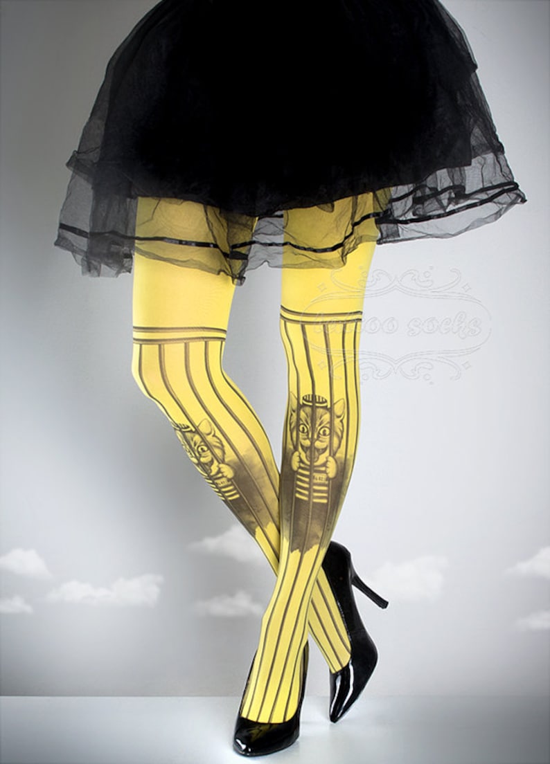 Puede incluir: Mallas amarillas con rayas negras y un estampado gr&aacute;fico de un gato con un gorro de marinero y una camisa a rayas. El gato est&aacute; detr&aacute;s de las rejas.