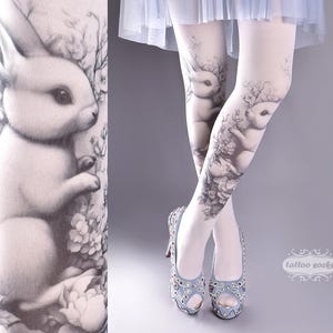 Könnte beinhalten: Weiße Strumpfhosen mit einem schwarz-weißen Kaninchen-Design. Das Design zeigt ein Kaninchen auf jedem Bein, umgeben von Blumen und Blättern. Die Strumpfhosen werden von einer Person in blauen Schuhen getragen.