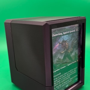 May include: A black card storage box with a clear front panel, displaying a Magic: The Gathering card. The card features the text "Chatterfang, Squirrel General" and a creature illustration. The box is designed to protect and showcase collectible cards.