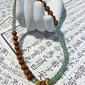 May include: A green and brown beaded bracelet with a gold ring charm. The beads are made of wood and jade. The bracelet is on a white surface.