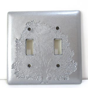 May include: Silver-colored double light switch cover with an embossed tree design. The cover has two rectangular switch openings and four screw holes. The tree design is centered and detailed.