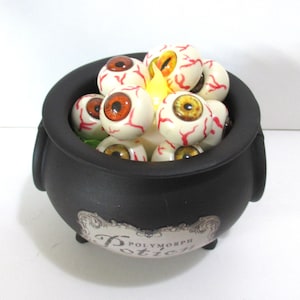 May include: A black cauldron with a label reading Polymorph Potion, filled with multiple realistic white eyeballs featuring red veins and various iris colors, illuminated by a small yellow light nestled among the spheres.
