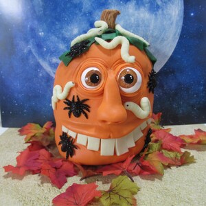 Könnte beinhalten: Ein Halloween-kürbiskuchen, der mit schwarzen Spinnen, weißen Würmern und Glitz Augen verziert ist. Der Kuchen ist orange und hat ein breites, zahniges Grinsen.
