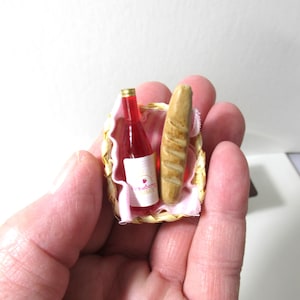 May include: A miniature wicker basket with a pink cloth liner contains a small bottle of strawberry wine and a miniature loaf of bread.