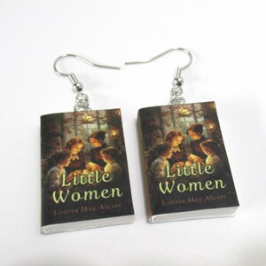 Op de afbeelding: Een paar oorhangers met miniatuur boekomslagen. Elke oorbel toont de omslag van "Little Women" van Louisa May Alcott, met een warmgekleurde illustratie van figuren binnenshuis. De oorbellen hebben zilverkleurige haken.