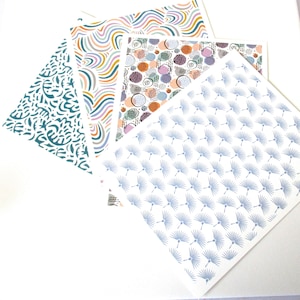 May include: Four patterned paper sheets arranged diagonally. The patterns include teal botanical, rainbow wave, orange and purple dot, and blue fan designs. The papers are on a white background.