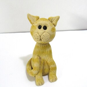 以下が含まれることがあります： 黒い目と質感のある表面を持つ、黄褐色と黄色の猫の置物。 猫は体を丸めて直立しています。 耳はピンと立ち、置物は気まぐれで手作りの外観をしています。