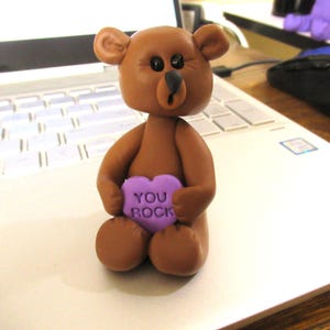 以下が含まれることがあります： 「YOU ROCK」の白い文字が入った紫色のハート型のキャンディーを持っている茶色のクマの置物。クマは黒い目と黒い鼻をしています。この置物は粘土または同様の素材で作られています。