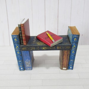 May include: A miniature bookshelf made of stacked books with a red book and a yellow pencil on top. The bookshelf is made of blue, brown, and black books.