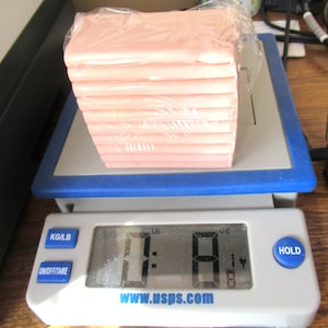 May include: A stack of pink rectangular items wrapped in clear plastic, resting on a digital scale. The scale displays a weight of 0.8 pounds. The scale has a blue border and the text "www.usps.com".