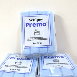 Może przedstawiać: Kilka opakowań jasno niebieskiej gliny do pieczenia Sculpey Premo. Każde opakowanie ma białą etykietę z nazwą marki, nazwą produktu i wagą 57 g. Etykieta zawiera również tekst w języku francuskim i hiszpańskim.