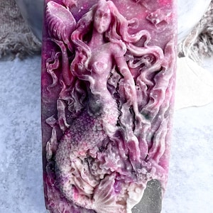 May include: A rectangular bar of soap with a carved mermaid design. The soap is primarily a deep pink color with lighter pink and white accents. The mermaid has flowing hair and a detailed tail, with fish and other aquatic elements.