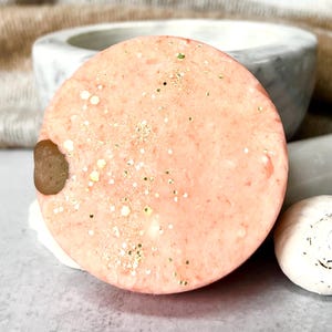 May include: A round, peach-colored soap bar with gold glitter embedded in the surface. The soap bar has a small, brown, irregular-shaped piece of soap attached to the side.