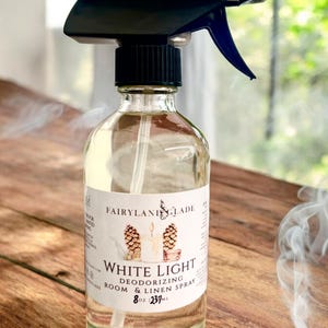May include: A clear glass bottle with a black spray top filled with a clear liquid. The bottle has a white label with the text "Fairyland Glade White Light Deodorizing Room & Linen Spray 8 oz | 237 ml".
