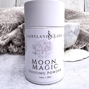 May include: A white cylindrical container of "Moon Magic Dusting Powder" with a floral moon design. The container has the text "Fairyland Glade" and "1 oz | 28g". The product is a cosmetic item.