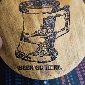 Op de afbeelding: Een houten onderzetter met een ingebrand ontwerp van een bierkan en de tekst "The Dwarf" en "BEER GO HERE."