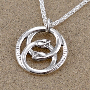 May include: Silver necklace with a circular pendant featuring two intertwined snakes with heads facing outward. The pendant has a textured border.