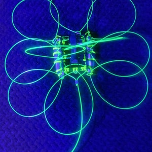 May include: A black drone with a green glow-in-the-dark cord attached to it. The cord is in the shape of a four-leaf clover.