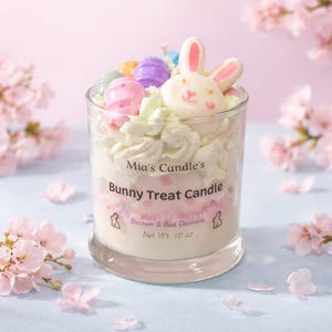 Puede incluir: Vela decorativa en un vaso transparente. La vela presenta un diseño de conejito y huevo, con un conejito blanco, huevos de colores pastel y una cobertura tipo crema batida. La etiqueta dice "Mia's Candle's Bunny Treat Candle", peso neto 283 gramos.