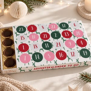 Pode incluir: Um calendário do Advento Toffifee retangular com fundo branco e padrão de renas verdes. A caixa exibe círculos numerados em vermelho, rosa e verde, com o texto ADVENTSKALENDER TO GO. Uma bandeja dourada contém quatro chocolates redondos.