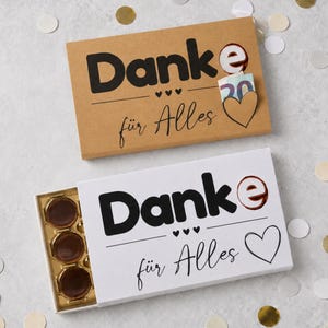 Può includere: Due scatole regalo rettangolari con il testo Danke für Alles e un'icona a forma di cuore. La scatola superiore è in cartone marrone con una banconota da venti euro inserita. La scatola inferiore è bianca e contiene tre cioccolatini rotondi in un vassoio dorato.