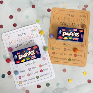 May include: Two rectangular cards with rounded corners, one white and one tan, each featuring the text "FÜR EIN KUNTERBUNTES SCHULJAHR" and "GANZ VIEL" with a list of attributes. Each card holds a small blue box of Smarties mini chocolate candies.