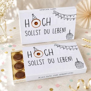 Könnte beinhalten: Eine weiße rechteckige Geschenkbox mit runden Pralinen in einem goldenen Einsatz. Der Deckel zeigt den schwarzen Schriftzug HOCH SOLLST DU LEBEN!, einen Partyhut, Wimpel und einen Cupcake. An der Seite steht ALLES GUTE ZUM GEBURTSTAG.