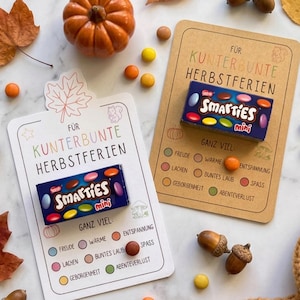 Puede incluir: Dos tarjetas rectangulares con esquinas redondeadas, cada una con una pequeña caja de caramelos Smarties mini adherida. Las tarjetas tienen el texto "FÜR KUNTERBUNTE HERBSTFERIEN" y una lista de palabras junto a círculos de colores, decoradas con dibujos.