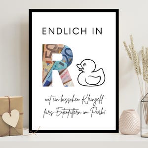 May include: A framed print with the words "ENDLICH IN" at the top. The letter "R" is filled with euro banknotes. A cartoon duck is to the right of the "R". The text at the bottom says "mit ein bisschen Kleingeld fürs Entenfüttern im Park!"
