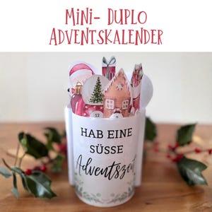 Könnte beinhalten: Ein weißer, becherförmiger Adventskalender mit dem Text "Mini-Duplo Adventskalender" in roter Schrift. Der Becher enthält weihnachtliche Papierauschnitte, darunter ein Weihnachtsmann, ein Weihnachtsbaum und ein Lebkuchenhaus. Der Becher trägt den Text "Hab eine süsse Adventszeit".