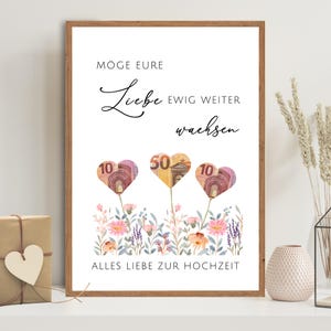 Könnte beinhalten: Ein gerahmter Druck mit dem Text "Möge eure Liebe ewig weiter wachsen" und "Alles Liebe zur Hochzeit". Das Kunstwerk zeigt herzförmige Blumen aus Euro-Banknoten mit einem floralen Rand. Der Rahmen ist braun.
