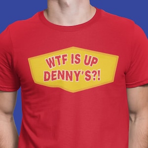 May include: A bright red short-sleeved crew neck t-shirt featuring a yellow hexagonal graphic on the chest. The graphic contains the red text "WTF IS UP DENNY'S?!" in a bold, sans-serif font, centered on the front of the garment.